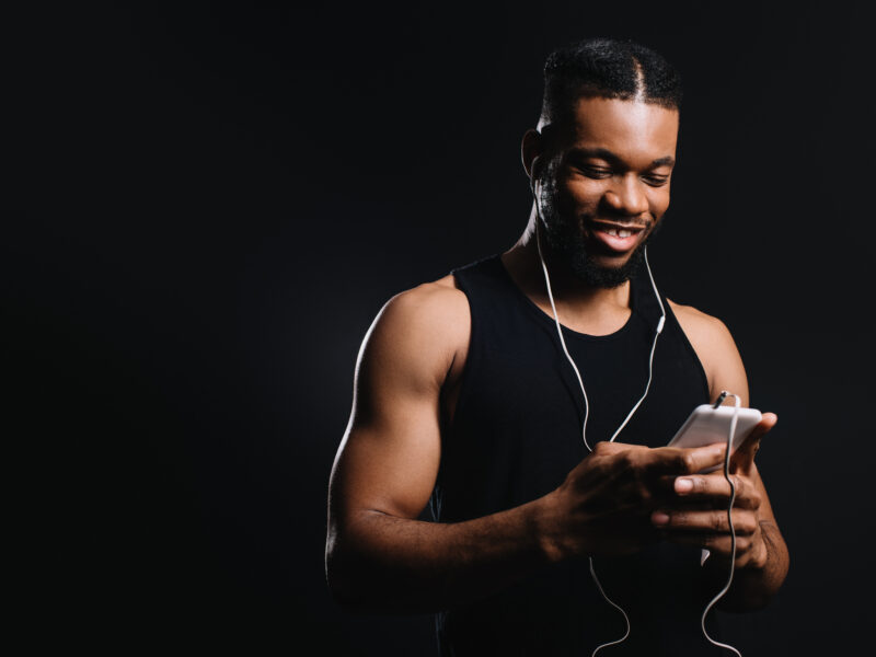 smiling muscular african american man in earphones using smartphone isolated on black